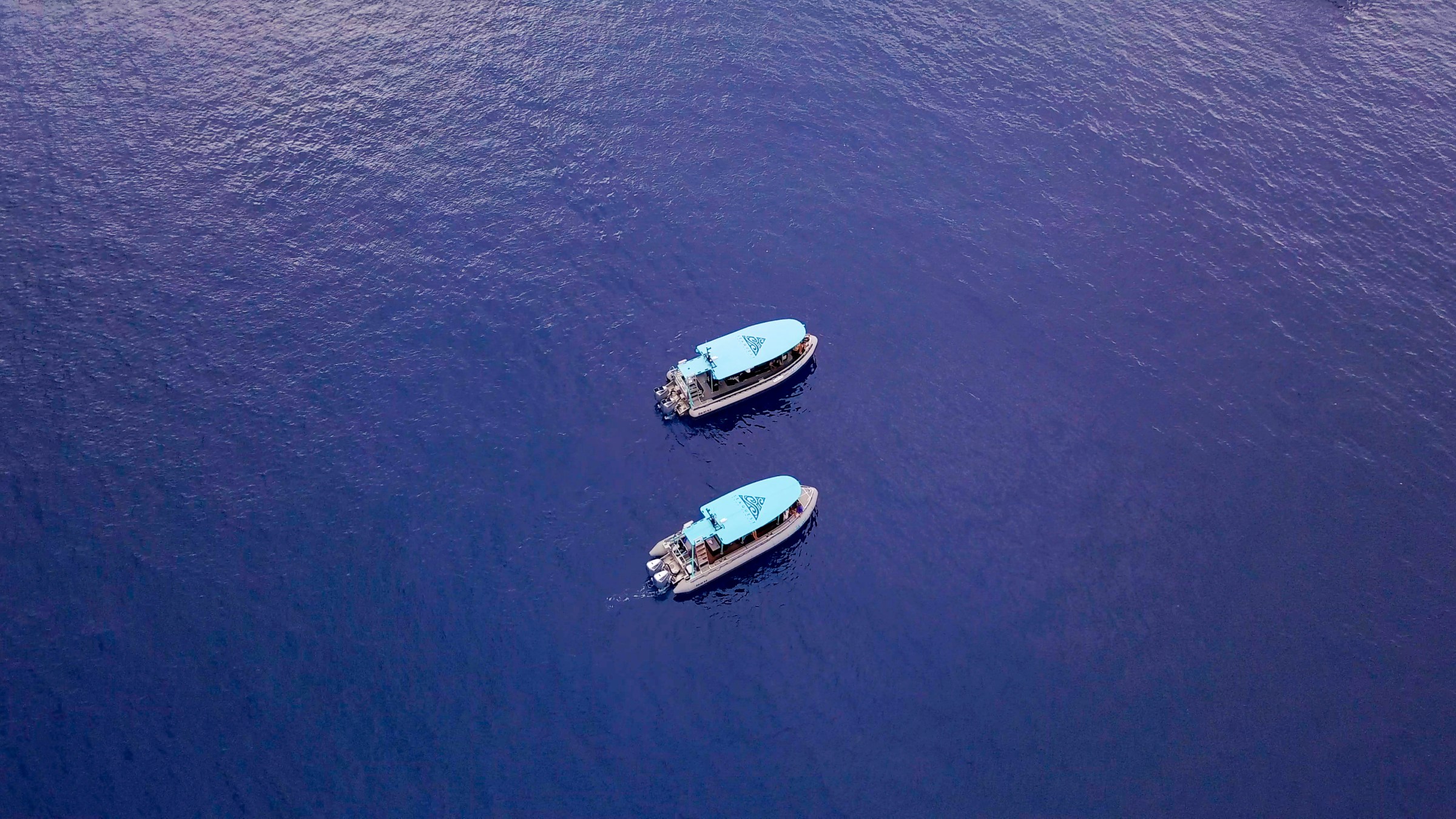 Two boats with blue canopies floating on calm, deep blue water, viewed from above.