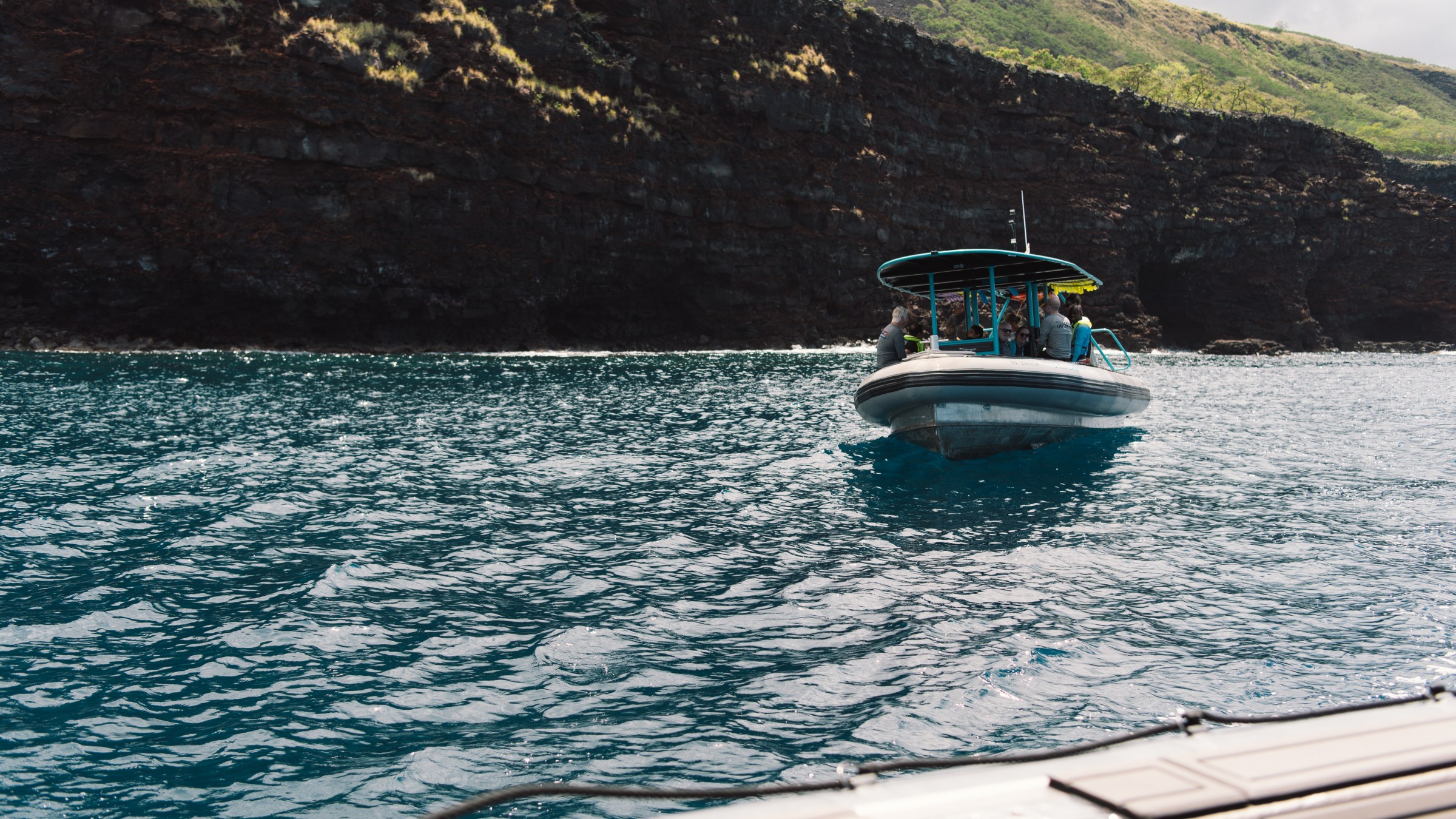 Small boat with people approaches a rocky cliff in blue ocean waters.