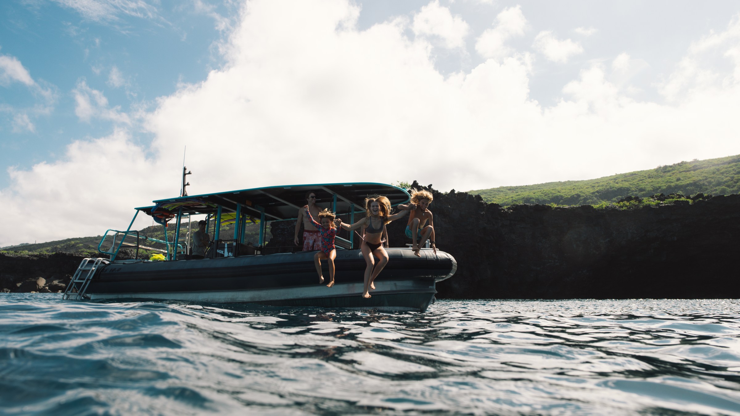 Three people jumping off a boat into the ocean, with cliffs and a cloudy sky in the background.