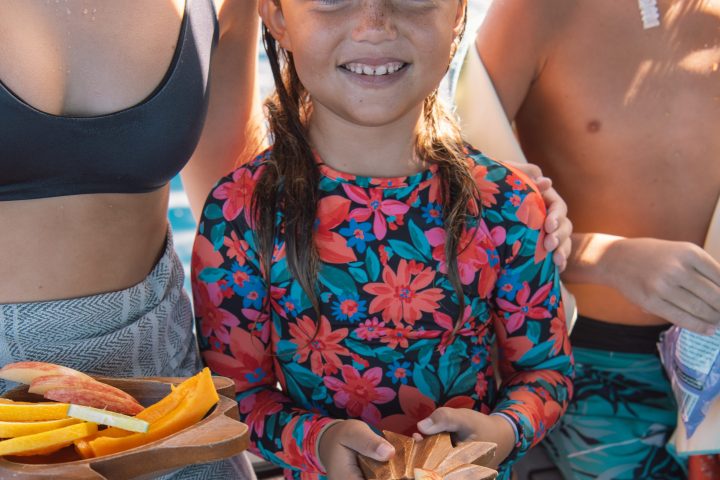 Child with floral swimsuit holding a wooden bowl of sliced fruit, standing between two people in swimwear.