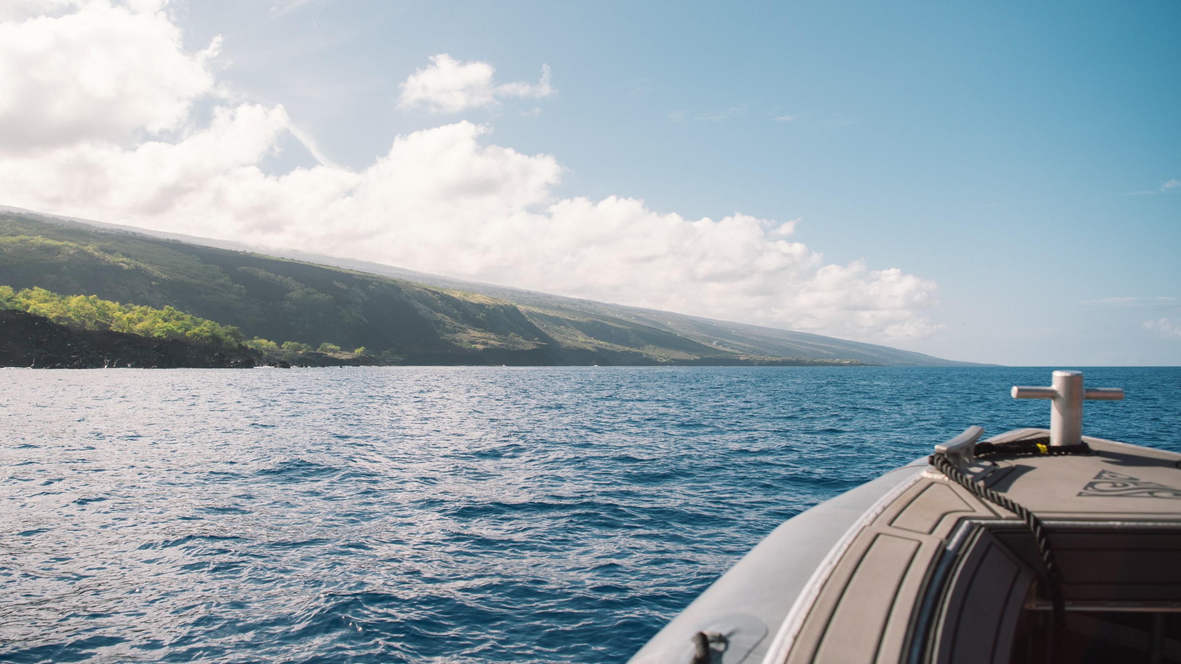 Boat on blue ocean near green hilly coast under a cloudy sky.