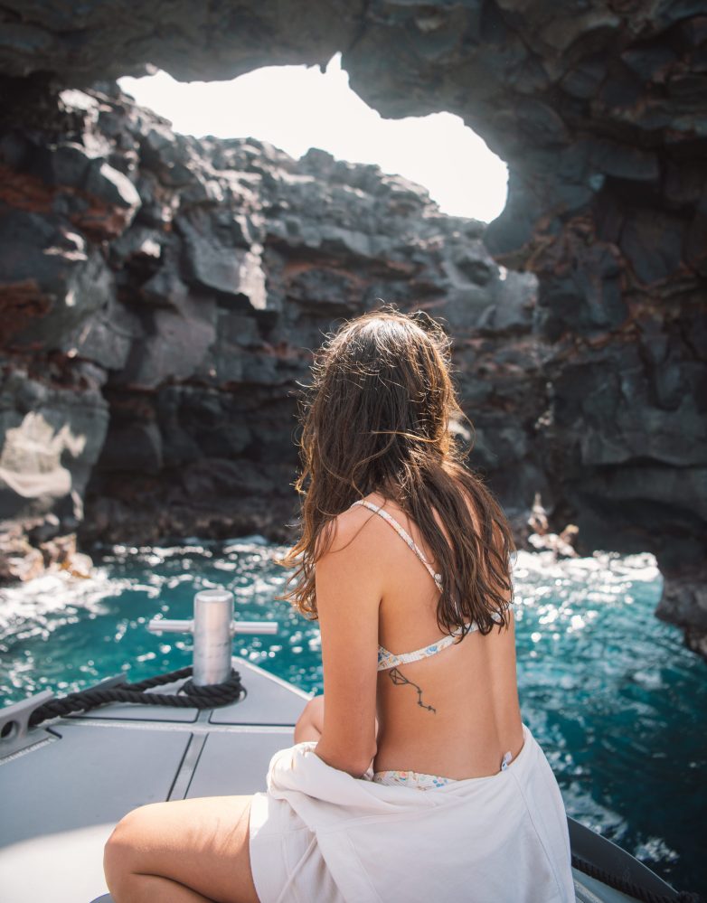 Woman in swimwear sits on a boat, looking at rocky sea cave opening.
