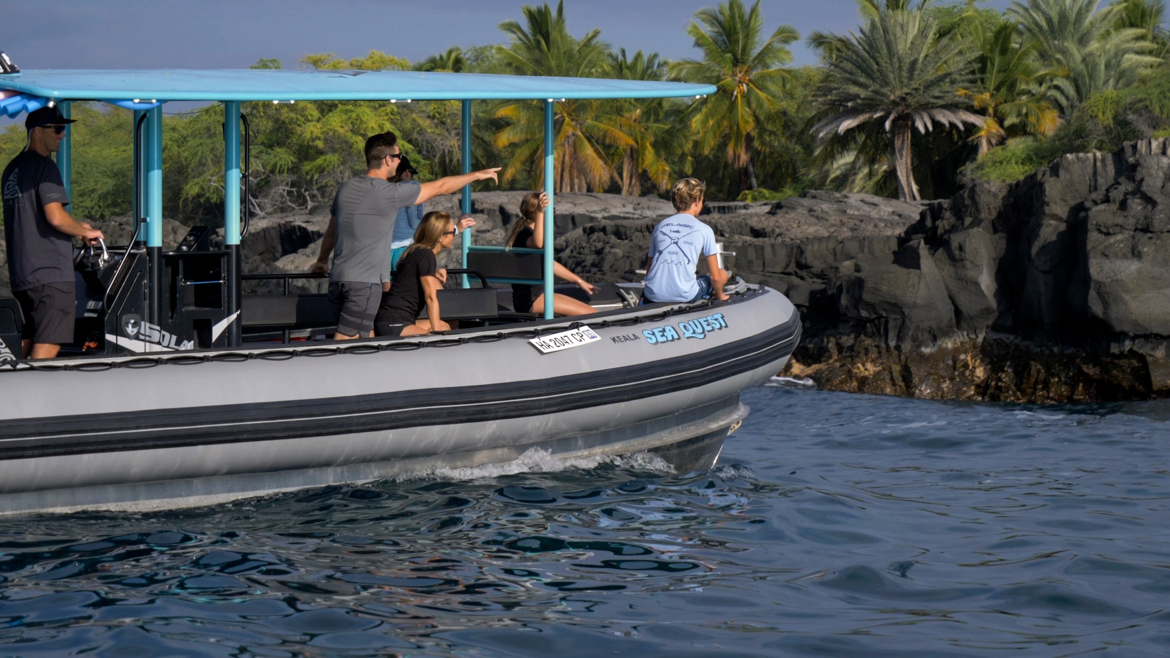 Group on a boat near rocky shore with palm trees in the background.