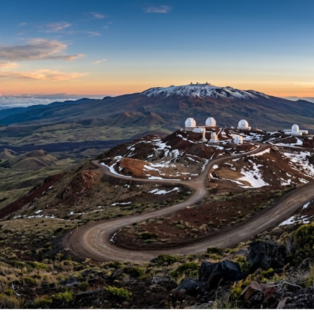 Telescopes on mountain summit with winding road and snowy patches, under a cloudy sky.