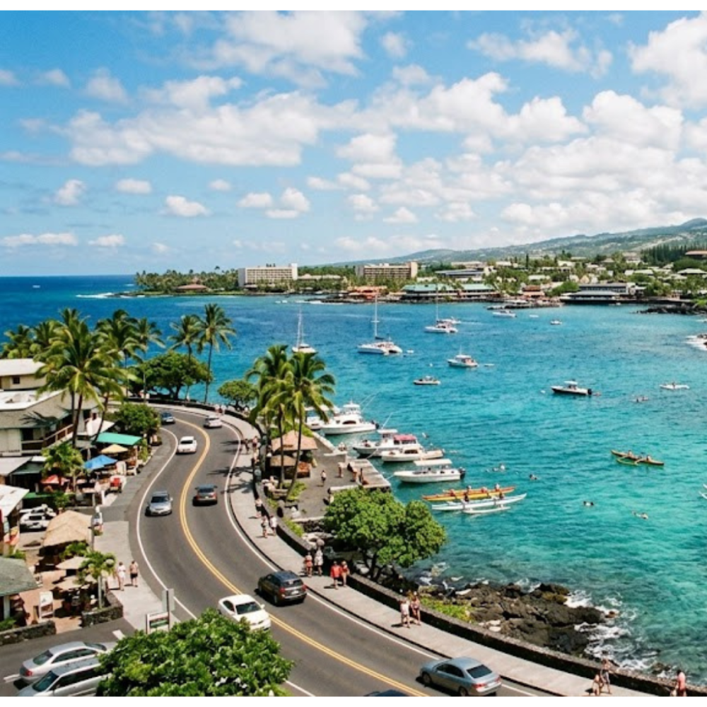 Coastal road with cars, tropical trees, and boats on blue water under a cloudy sky.