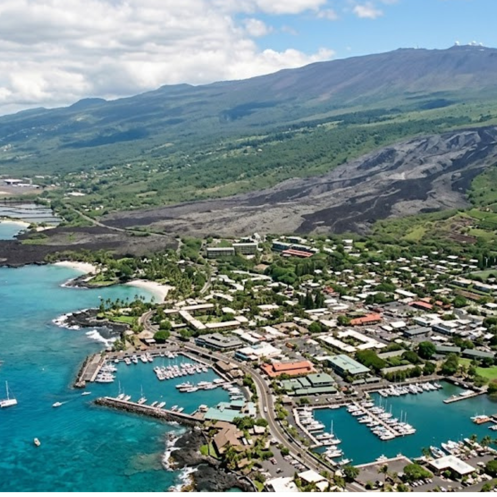 Aerial view of a coastal town with a marina and lush green mountain backdrop.