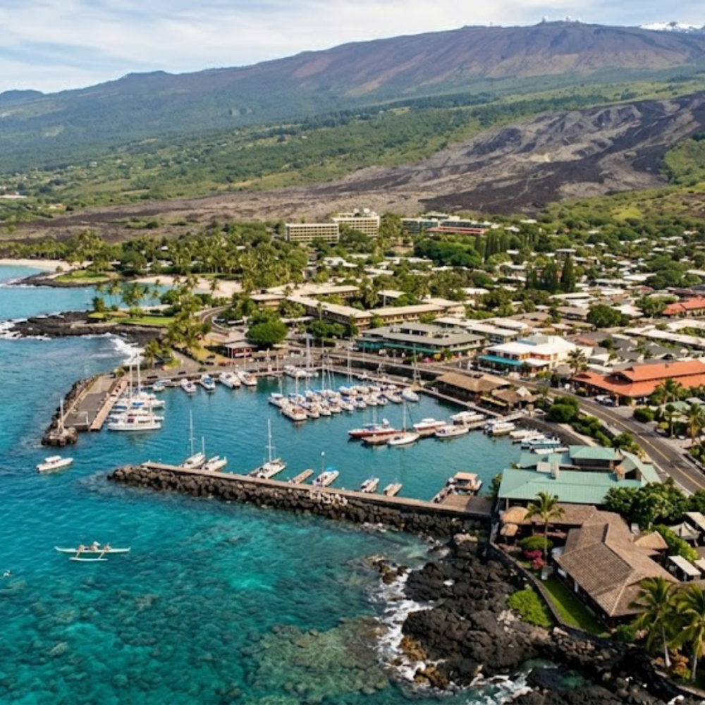 Coastal town with a marina, boats, and lush green hills in the background.